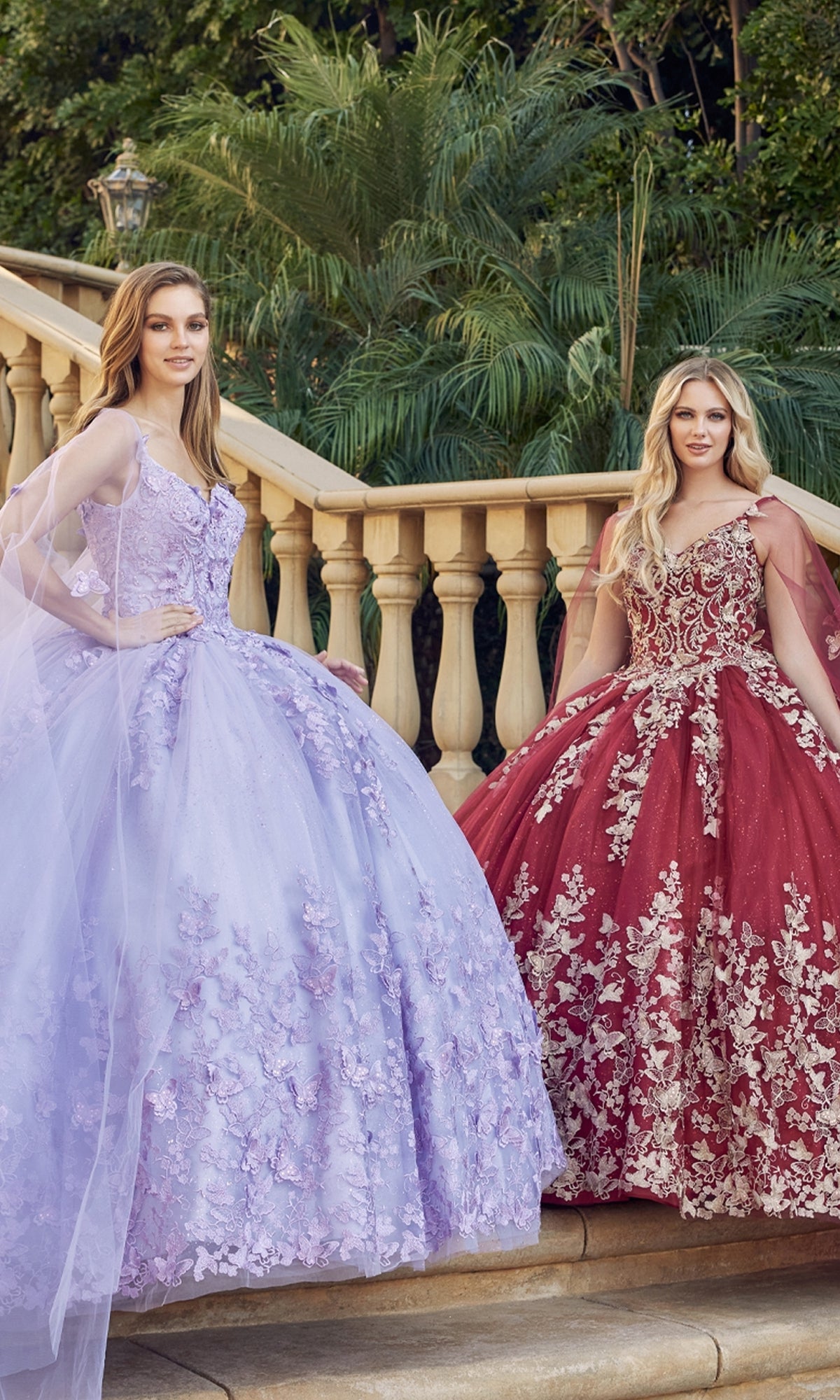 Two women in Juliet 1442 Quinceañera Dresses with Sheer Capes stand on outdoor stone steps; one wears lavender, the other burgundy. Both gowns have floral embroidery and voluminous skirts.